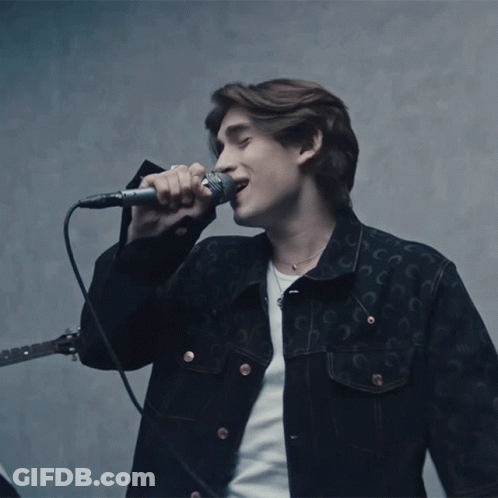 A Man Singing Into A Microphone While Wearing A Black Jacket With The Letter C On It GIF