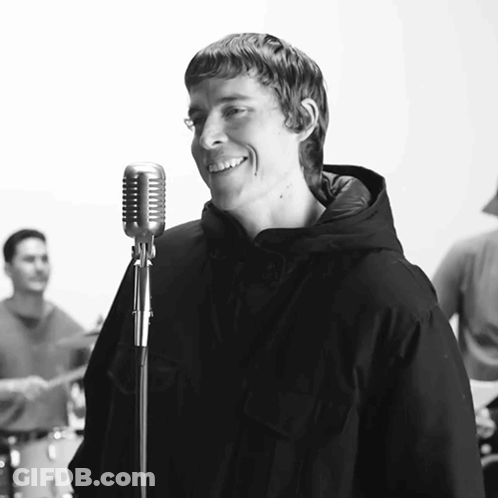 A Man Singing Into A Microphone With A Band Behind Him GIF