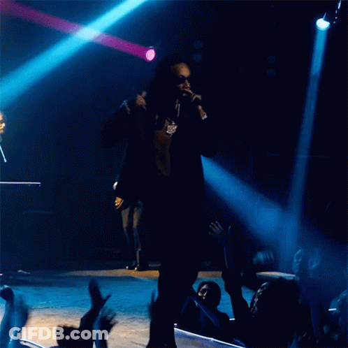 A Man Singing Into A Microphone With A Crowd Behind Him GIF