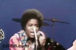 A Man Singing Into A Microphone With A Drum Set Behind Him GIF