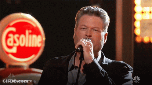 A Man Singing Into A Microphone With A Gasoline Sign In The Background GIF