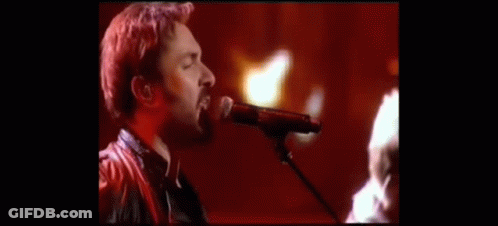 A Man Singing Into A Microphone With A Guitar In The Background GIF