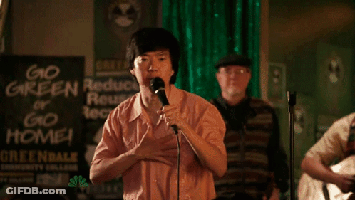 A Man Singing Into A Microphone With A Sign Behind Him That Says Go Green Or Go Home GIF