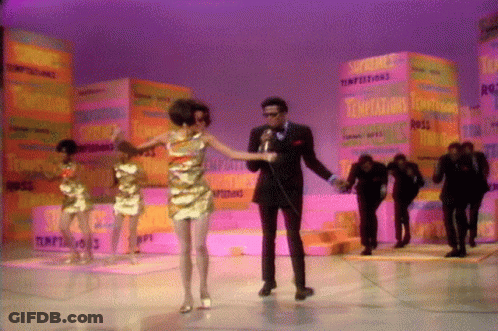 A Man Singing Into A Microphone With A Woman Dancing In Front Of A Stack Of Boxes That Say Temptations GIF