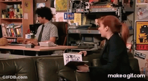 A Man Sits At A Desk While A Woman Sits On A Couch With A Book In Her Hand GIF