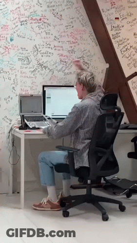 A Man Sits At A Desk With A Laptop In Front Of A Wall That Has Graffiti On It Including The Name Camelle GIF