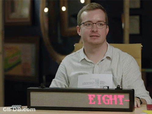 A Man Sits At A Desk With A Sign That Says Eight On It GIF