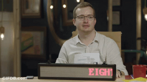 A Man Sits At A Desk With A Sign That Says Eight On It GIF