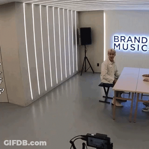 A Man Sits At A Table In A Room With A Sign That Says Brand Music GIF