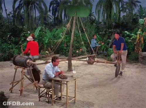 A Man Sits At A Table While Another Man Rides A Wheelbarrow GIF