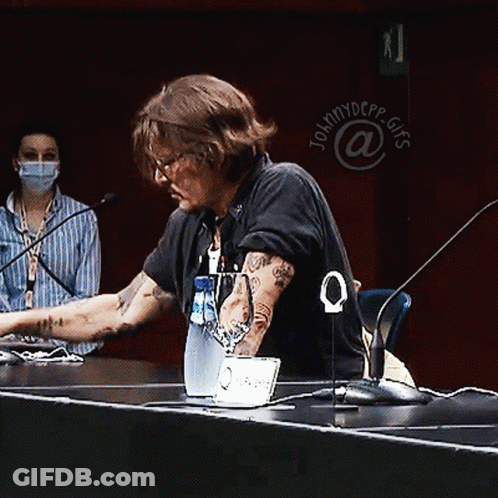 A Man Sits At A Table With A Laptop And A Bottle Of Water In Front Of A Sign That Says Johnny Depp GIF