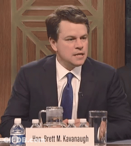 A Man Sits At A Table With A Sign That Says Hon. Brett M. Kavanugh GIF