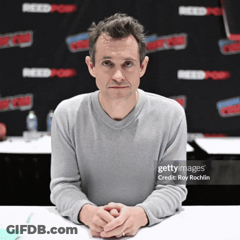 A Man Sits At A Table With His Hands Folded In Front Of A Wall That Says Reedpop GIF