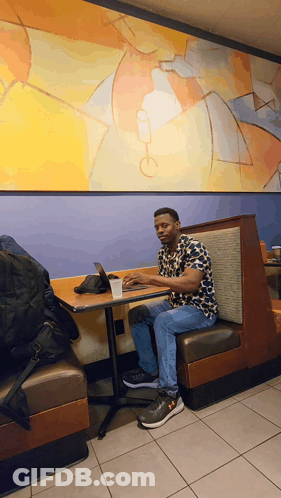A Man Sits In A Booth At A Table With A Laptop On It GIF