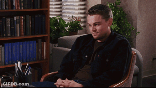 A Man Sits In A Chair In Front Of A Bookshelf With A Blue Book Titled " A Brief History Of The World " GIF
