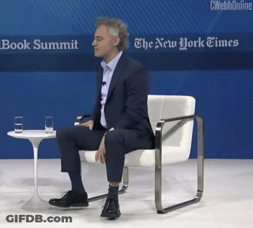 A Man Sits In A Chair In Front Of A Sign That Says The New York Times Book Summit GIF
