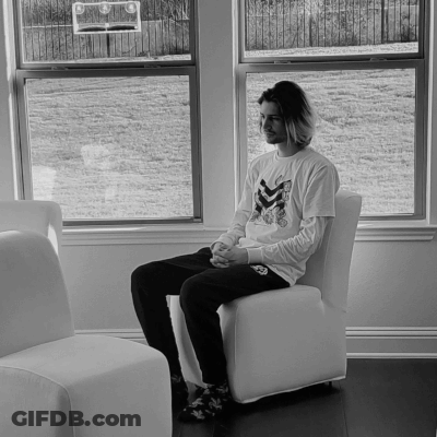 A Man Sits In A Chair In Front Of A Window Wearing A White Shirt With A Skeleton On It GIF