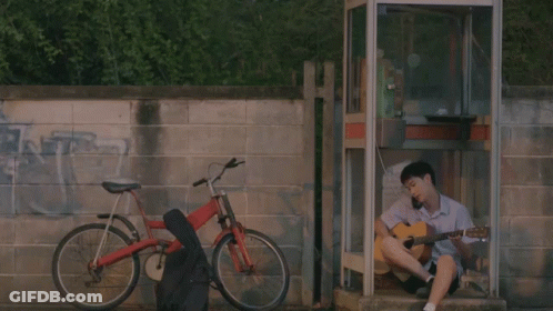A Man Sits In A Phone Booth Playing A Guitar Next To A Bike GIF
