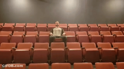 A Man Sits In A Row Of Red Seats In A Theater GIF