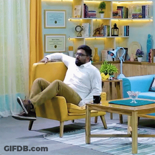 A Man Sits In A Yellow Chair In A Living Room With A Diploma On The Wall Behind Him GIF