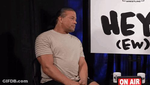 A Man Sits In Front Of A Sign That Says Hey GIF