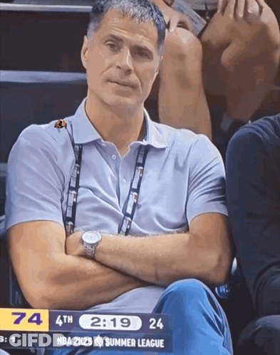 A Man Sits In The Stands With His Arms Crossed And A Nba 2K25 Summer League Scoreboard In The Background GIF