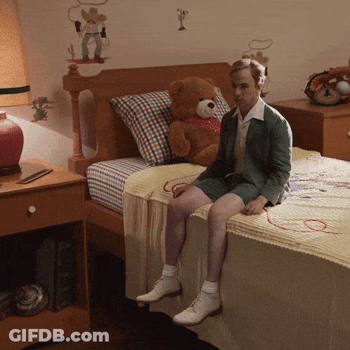A Man Sits On A Bed With A Teddy Bear On It GIF