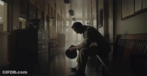 A Man Sits On A Bench In A Hallway Holding His Hat GIF