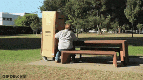 A Man Sits On A Bench In A Park While Carrying A Box GIF
