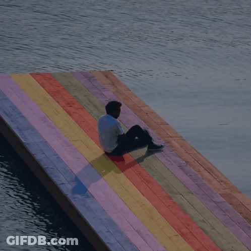 A Man Sits On A Colorful Wooden Dock Overlooking A Body Of Water GIF