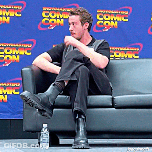 A Man Sits On A Couch In Front Of A Blue Wall That Says Showmasters Comic Con GIF