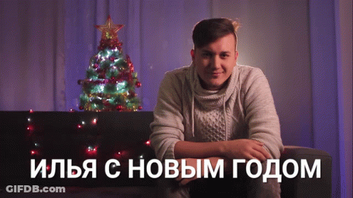 A Man Sits On A Couch In Front Of A Christmas Tree With Russian Writing On The Bottom GIF