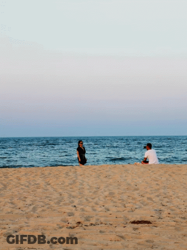 A Man Sits On The Beach While A Woman Stands On The Sand GIF