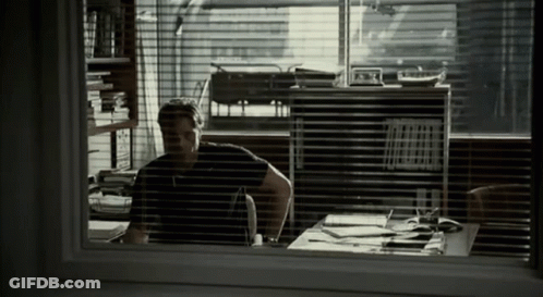 A Man Sitting At A Desk In Front Of A Window With Blinds GIF