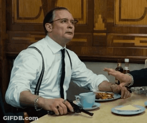 A Man Sitting At A Table Holding A Gun In His Hand GIF