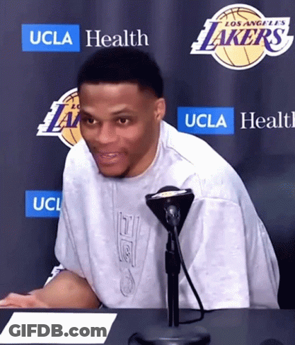 A Man Sitting At A Table In Front Of A Los Angeles Lakers Sign GIF