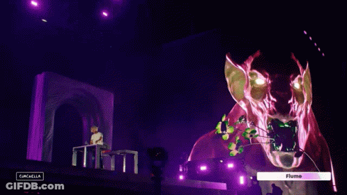 A Man Sitting At A Table In Front Of A Stage With A Sign That Says " Flume " On It GIF