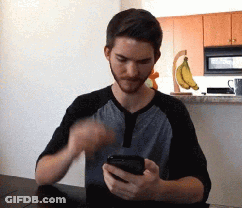 A Man Sitting At A Table Looking At A Cell Phone GIF
