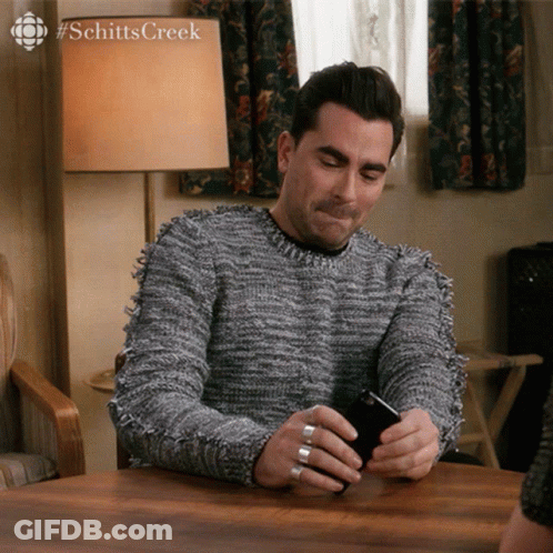 A Man Sitting At A Table Looking At A Cell Phone With #Schittscreek Written On The Bottom GIF