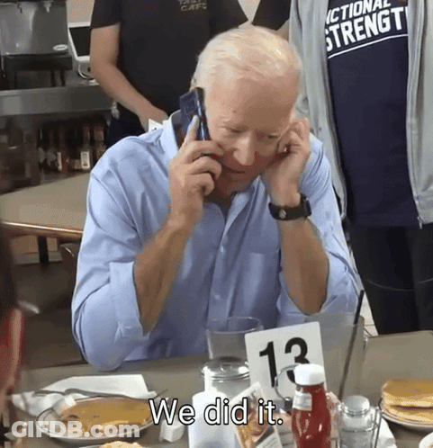 A Man Sitting At A Table Talking On A Cell Phone With A Sign That Says We Did It GIF