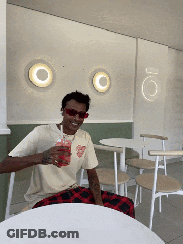 A Man Sitting At A Table With A Drink In His Hand GIF