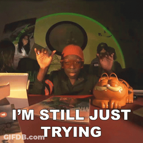 A Man Sitting At A Table With A Garfield Figurine And The Words I 'M Still Just Trying GIF