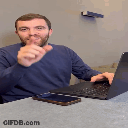 A Man Sitting At A Table With A Laptop And Cell Phone GIF