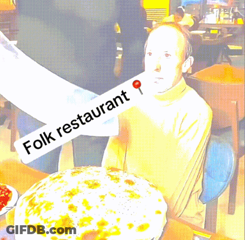 A Man Sitting At A Table With A Large Pizza And A Sign That Says Folk Restaurant GIF