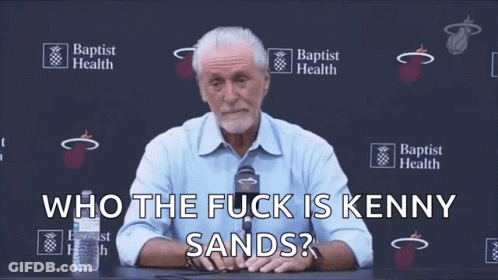 A Man Sitting At A Table With A Microphone And The Words Who The Fuck Is Kenny Sands Behind Him GIF