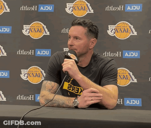 A Man Sitting At A Table With A Microphone In Front Of A Wall That Says Ajdu GIF