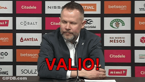 A Man Sitting At A Table With A Microphone In Front Of A Wall With Logos For Various Companies Including Valio GIF