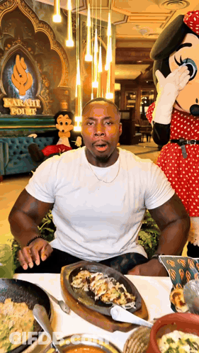 A Man Sitting At A Table With A Minnie Mouse Mascot Covering His Mouth GIF