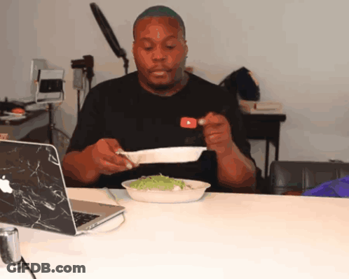 A Man Sitting At A Table With A Plate Of Food In Front Of A Laptop GIF