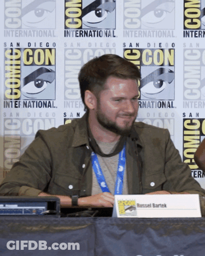 A Man Sitting At A Table With A Sign That Says Russell Bartek GIF